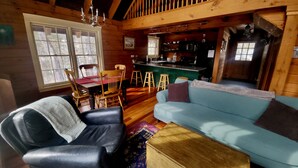 Interior acolhedor de cabana com sala de jantar, sala de estar com sofá azul, um baú de madeira e área de cozinha com balcão.