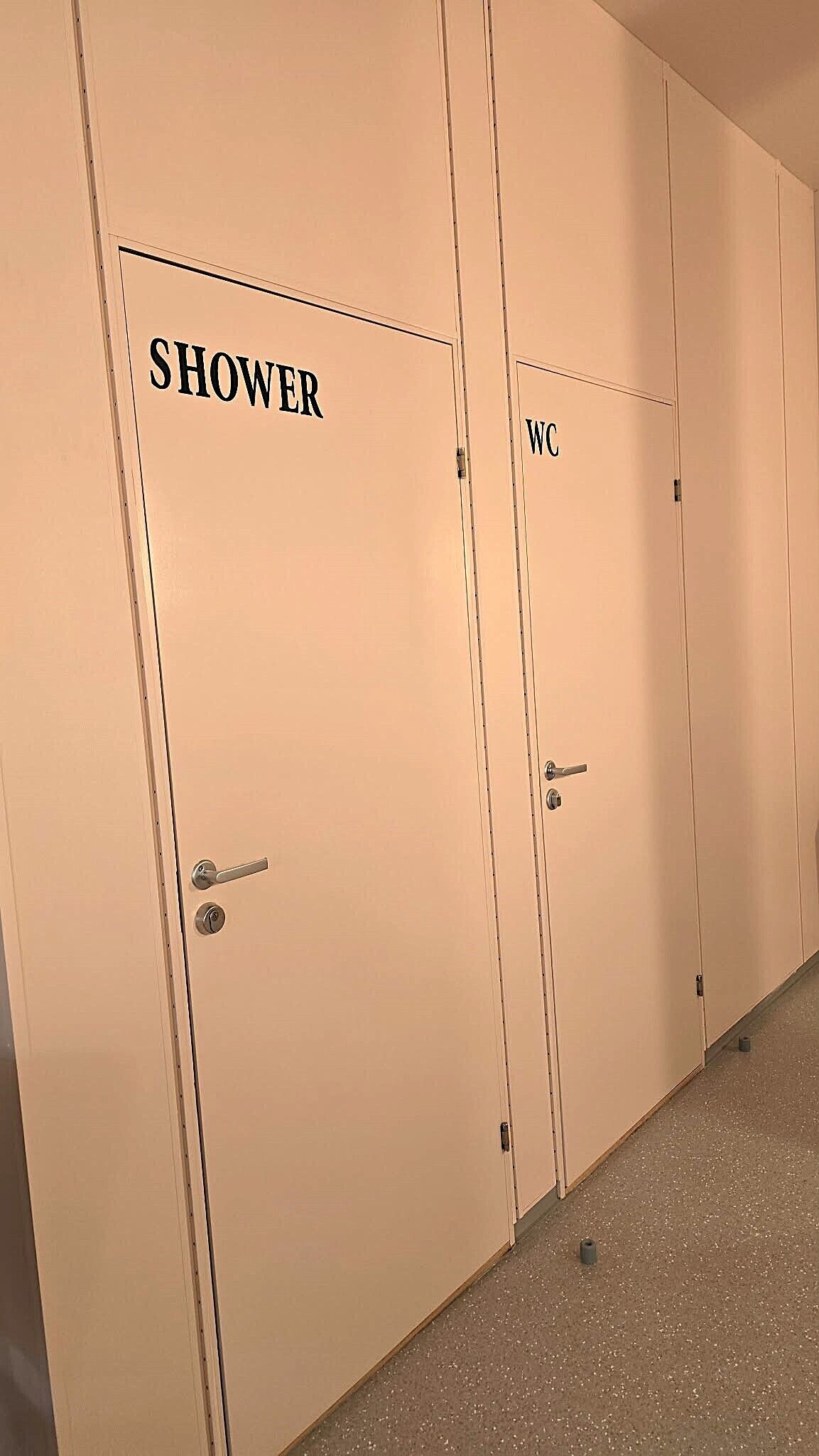 Egy sor bézs színű ajtó, melyeken a „ZUHANYZÓ” és a „WC” feliratok jelzik a fürdőszoba területét.