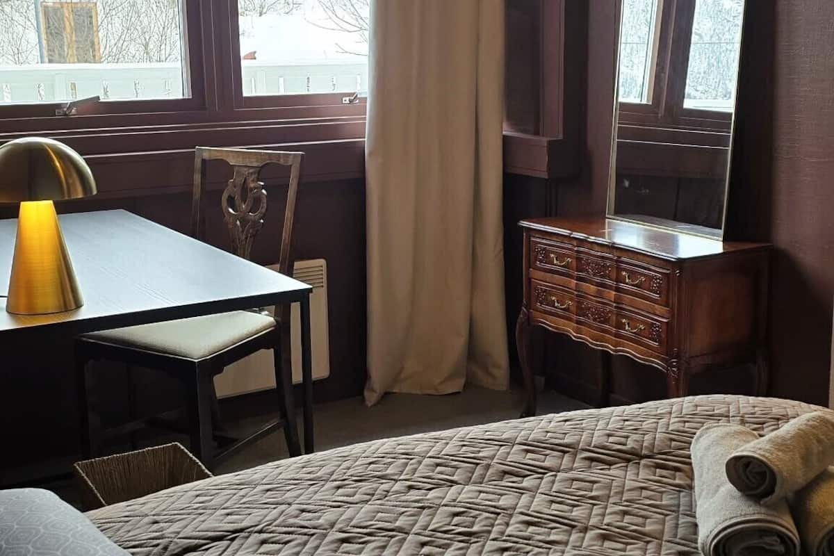 A hotel room with a bed, a desk, a chair, a dresser, and a large window with curtains.