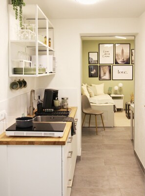 Une cuisine compacte équipée d’une cuisinière, d’un évier et d’étagères où sont rangés la vaisselle et les verres. On aperçoit à l’arrière-plan un coin repas avec une chaise et une table.