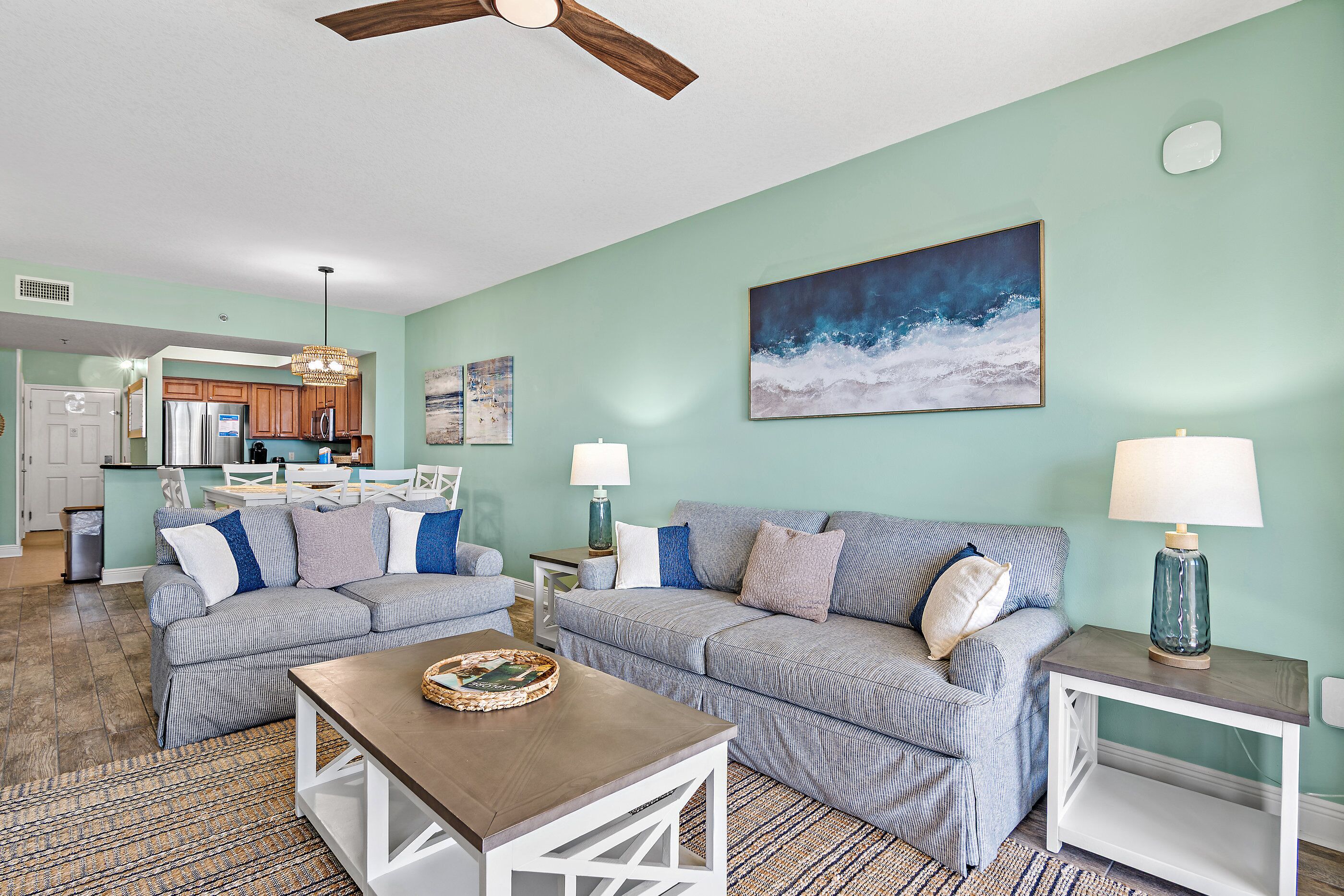 A living room with a sofa, coffee table, and lamps. There is a kitchen in the background.
