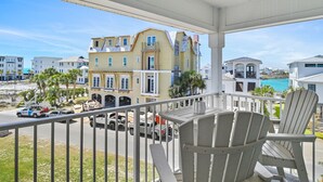 Uma varanda com cadeiras Adirondack brancas, vista para uma praia e um edifício com vários andares e varandas.