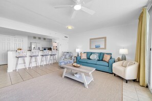 A modern living room with a blue sofa, white armchairs, a coffee table, and a kitchen area in the background. - 🌴Gulf Front with Amazing Views | Awesome Amenities | Emerald Beach 1928🌴 (Panama City Beach)