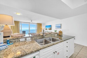 A kitchen with granite countertops, white cabinets, a double sink, and a view of the ocean through large windows. - 🌴Gulf Front with Amazing Views | Awesome Amenities | Emerald Beach 1928🌴 (Panama City Beach)