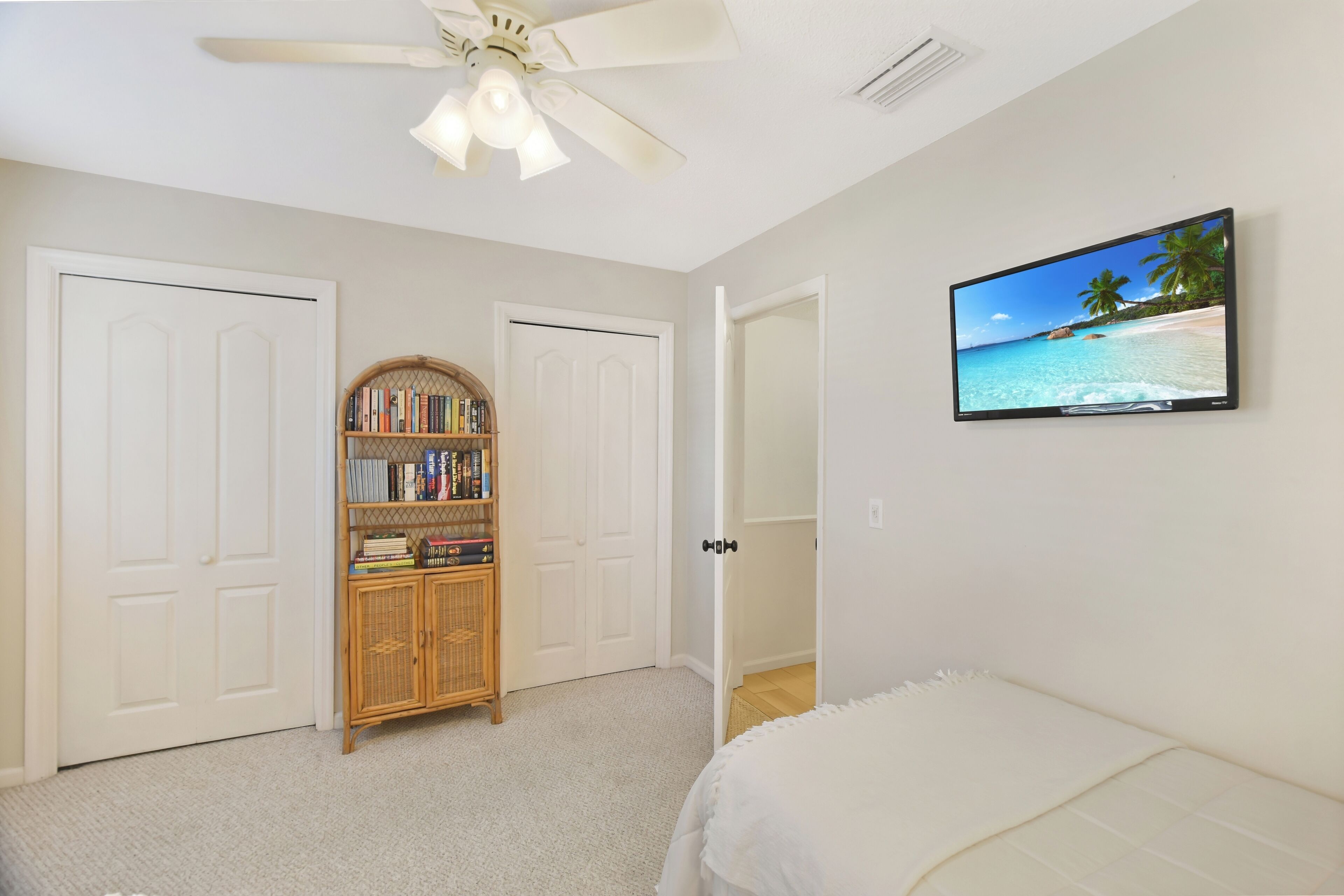 A bedroom with a bed, a ceiling fan, a bookshelf, and a TV displaying a beach scene.