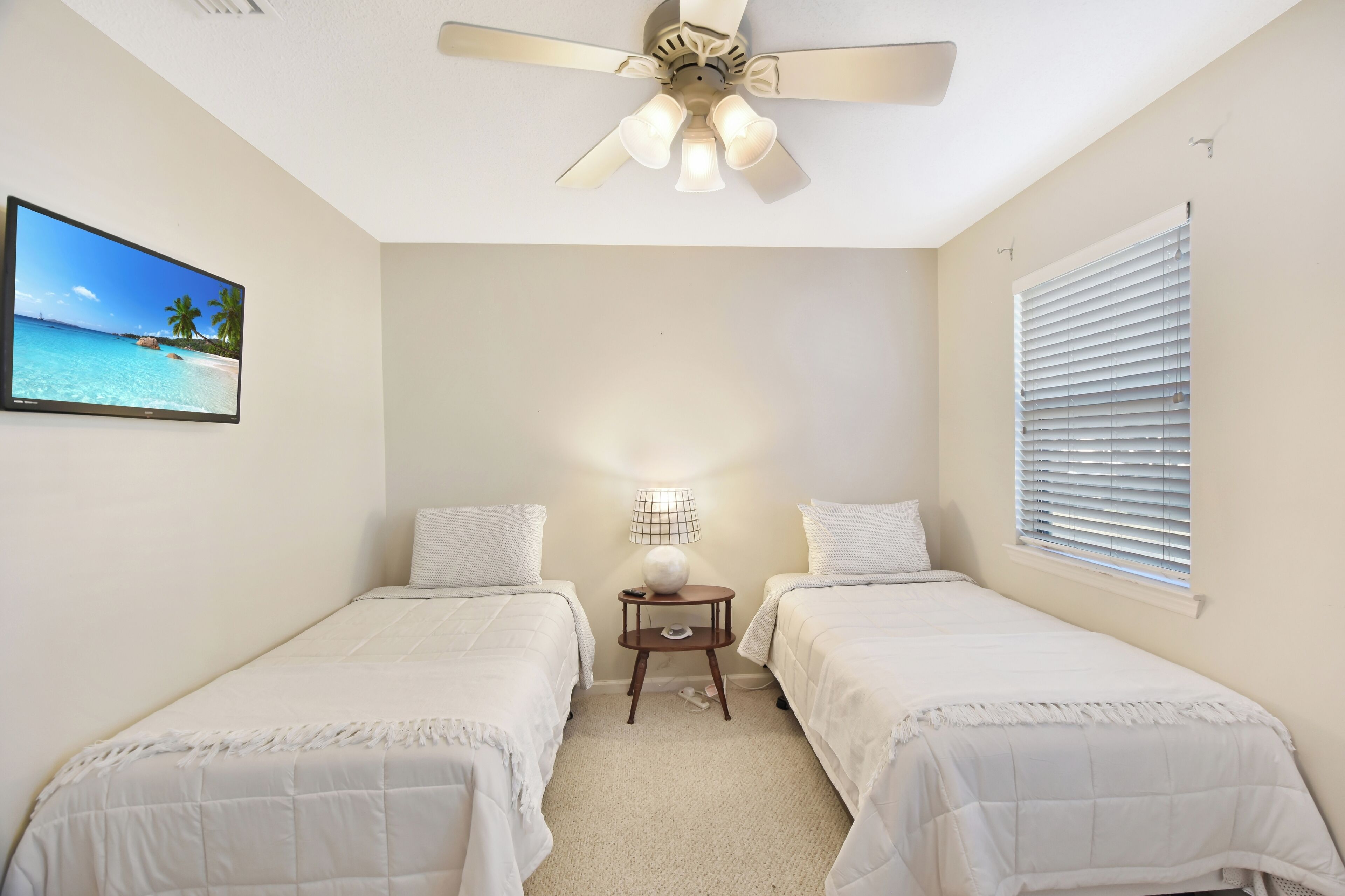 Une chambre avec deux lits, un ventilateur de plafond, un téléviseur fixé au mur, une lampe et une fenêtre avec des stores.