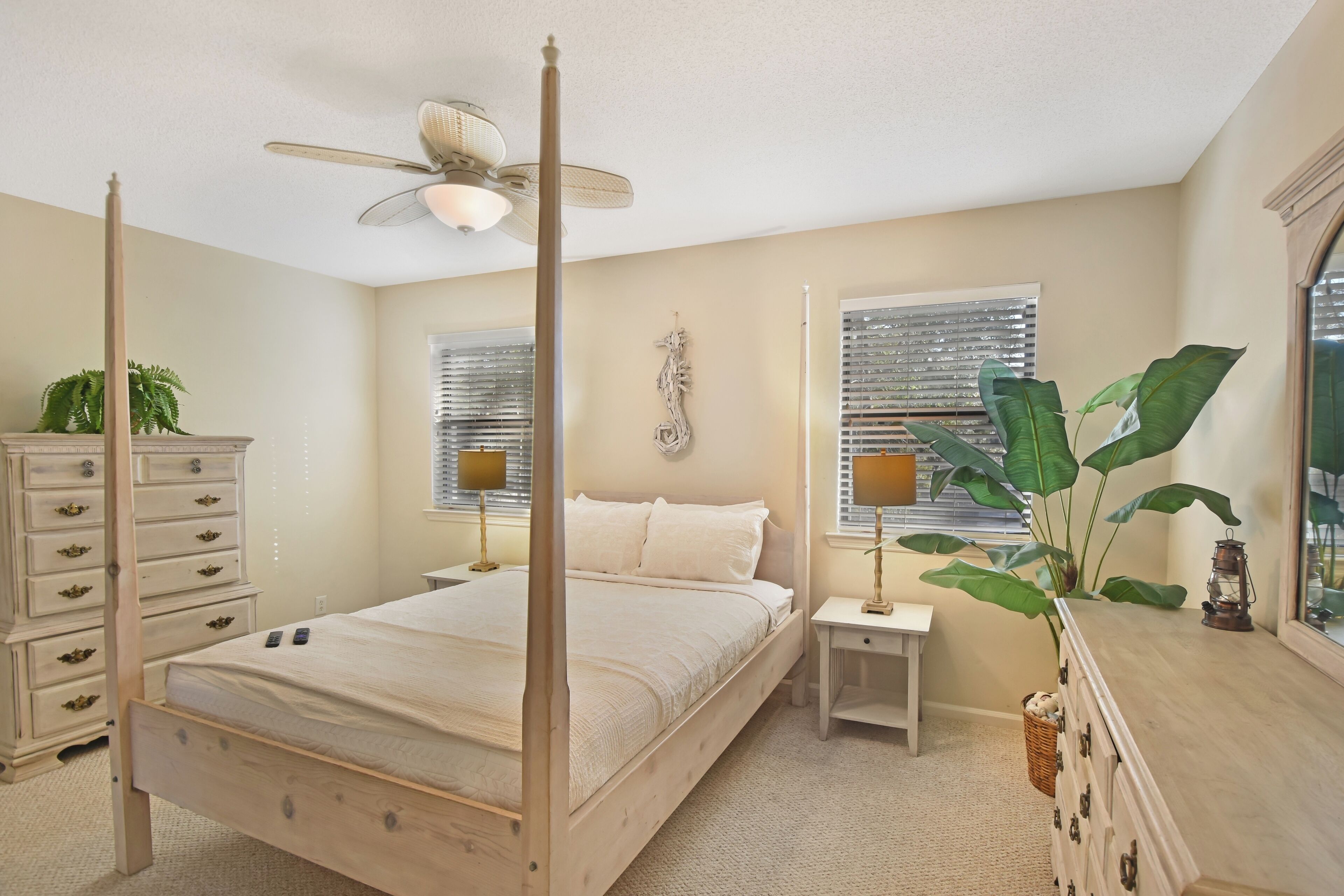 A four-poster bed with a ceiling fan, bedside tables with lamps, a dresser, and a large plant.