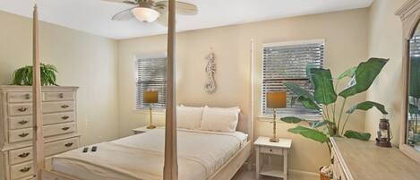 A four-poster bed with a ceiling fan, bedside tables with lamps, a dresser, and a large plant.