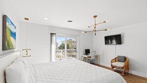Un dormitorio moderno con una cama grande, un escritorio, una silla, un televisor y un balcón con vistas a edificios y vegetación.