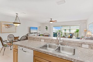 A kitchen with a double sink, wooden cabinets, and a dining area with wicker chairs. - Captain’s Cove 305 | Family-Friendly Condo w/ Pool, Dock & Easy Beach Access (Indian Shores)