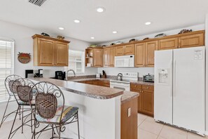 A modern kitchen with wooden cabinets, a white refrigerator, and a built-in island with bar stools. - Captain’s Cove 305 | Family-Friendly Condo w/ Pool, Dock & Easy Beach Access (Indian Shores)