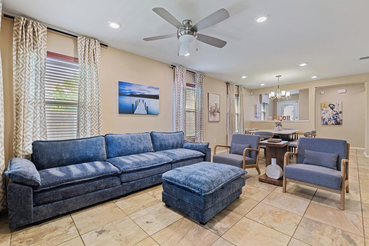 A living room with a sofa, ottoman, and chairs, a dining area with a table and chairs, a ceiling fan, and artwork on the walls.