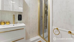 Un baño con tocador blanco, detalles dorados y ducha con mampara de vidrio.