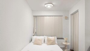 Un dormitorio con cama, almohadas, una mesita de noche con lámpara, un radiador y una ventana con persianas verticales.