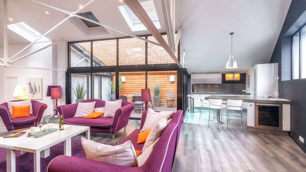 Una sala de estar moderna con sofás morados, una mesa de centro blanca y una cocina con una isla grande y taburetes de bar.