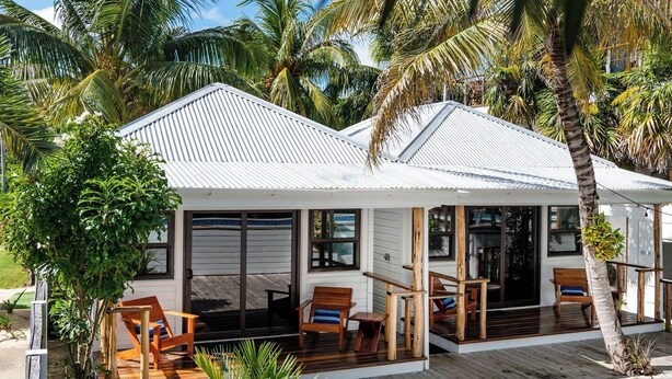 A two-story house with a covered porch, wooden furniture, and palm trees.