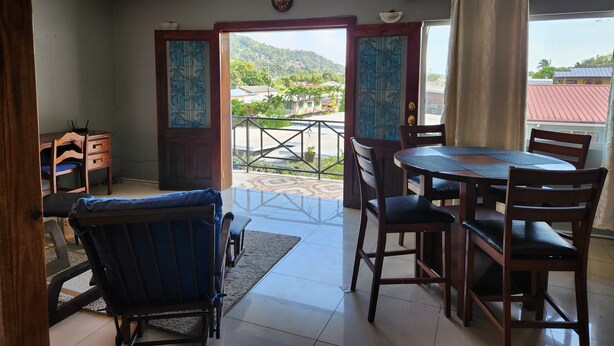 Un coin repas avec une table et des chaises, une vue sur un paysage montagneux par des portes ouvertes, et une horloge au mur.