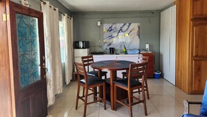 Un coin repas avec une table et des chaises en bois, une cuisine équipée d’un four à micro-ondes et d’une bouilloire, et un grand tableau abstrait accroché au mur.