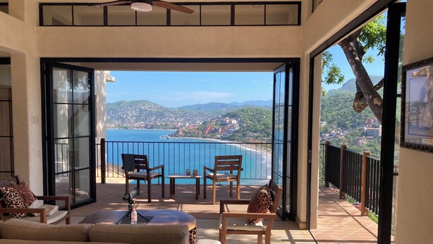 A balcony with a view of the sea and hills, furnished with chairs and a table, and a ceiling fan. - Playa La Ropa 
Brand new Villa with 7-minute walk to the beach (Zihuatanejo)