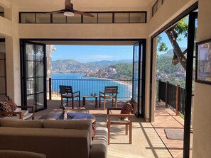 Uma varanda com vista para o mar e colinas, mobiliada com cadeiras e uma mesa, e um ventilador de teto.