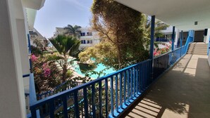 En balkon med blåt gelænder, udsigt over et poolområde med forskellige planter og en bygning i baggrunden.