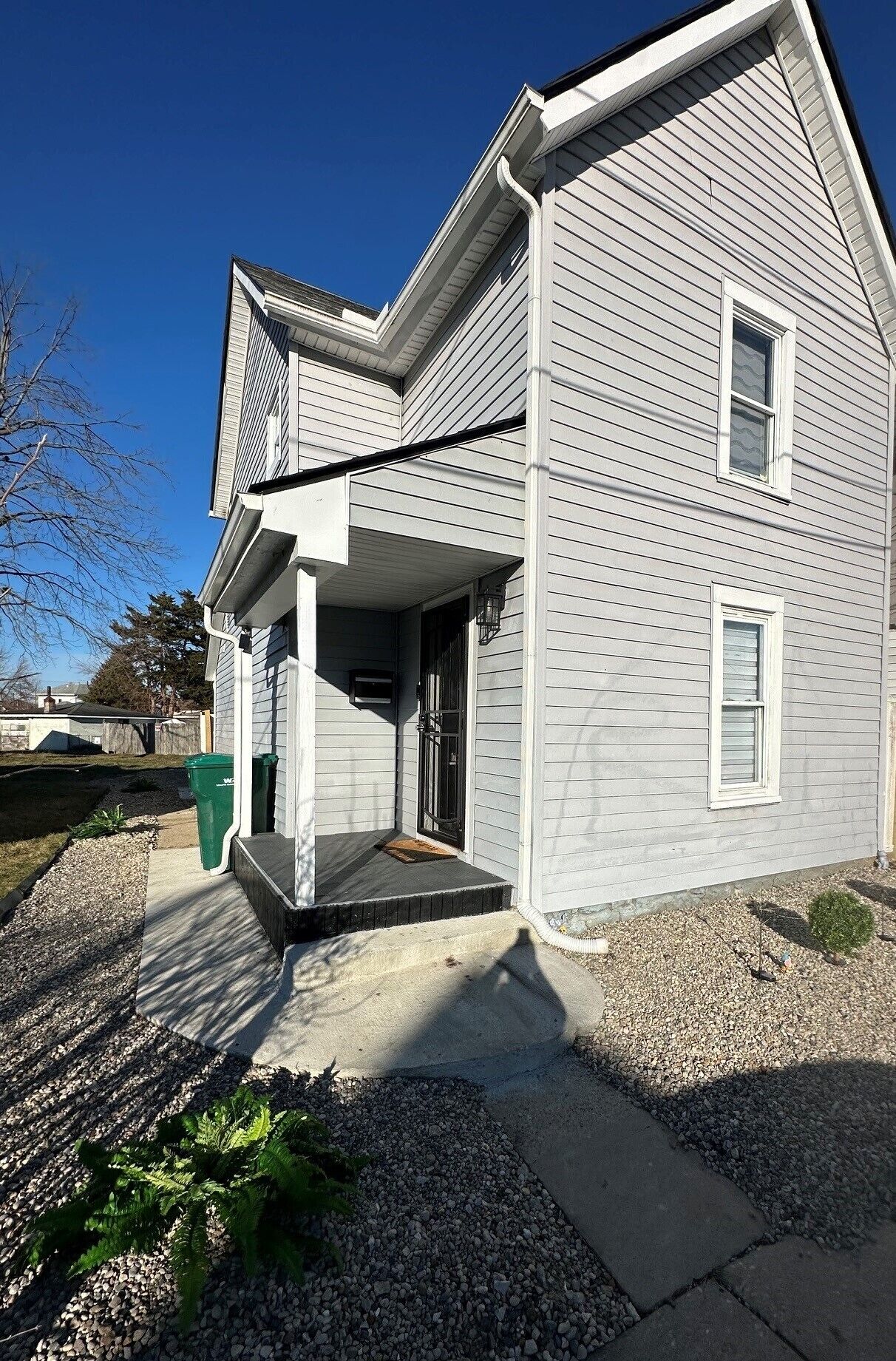 Une maison à deux étages avec un porche couvert, un petit jardin devant la maison avec du gravier et une poubelle verte.