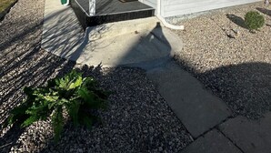 Een tweelaags huis met een overdekte veranda, een klein voortuintje met grind en een groene vuilnisbak.