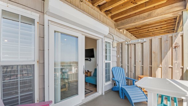 A wooden deck with a white table and colorful chairs, a sliding glass door, and a sign reading "SEASIDE".