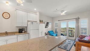 A modern kitchen with white cabinets, a granite countertop, and a built-in coffee maker. There is a blue chair and a wooden bench in the living area, and a balcony with a view.