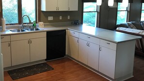 Una cocina moderna con armarios blancos, lavavajillas y una isla grande. Hay tres lámparas colgantes y una ventana con vistas a zonas verdes.