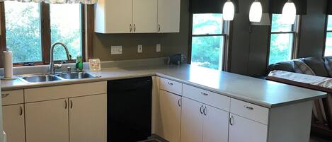 Une cuisine moderne avec des meubles blancs, un lave-vaisselle et un grand îlot central. On y trouve trois suspensions et une fenêtre donnant sur un espace verdoyant.