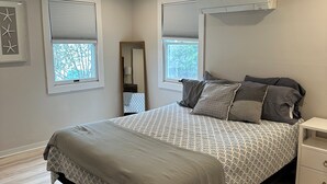 A bedroom with a large bed, two windows with blinds, a ceiling fan, and a nightstand.