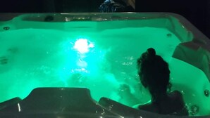 Un jacuzzi con una persona adentro, rodeado de plantas y un muro de madera con luces.