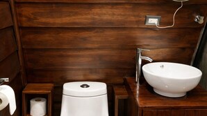 Un baño de madera con inodoro blanco, lavamanos, espejo y un pequeño estante con papel higiénico.