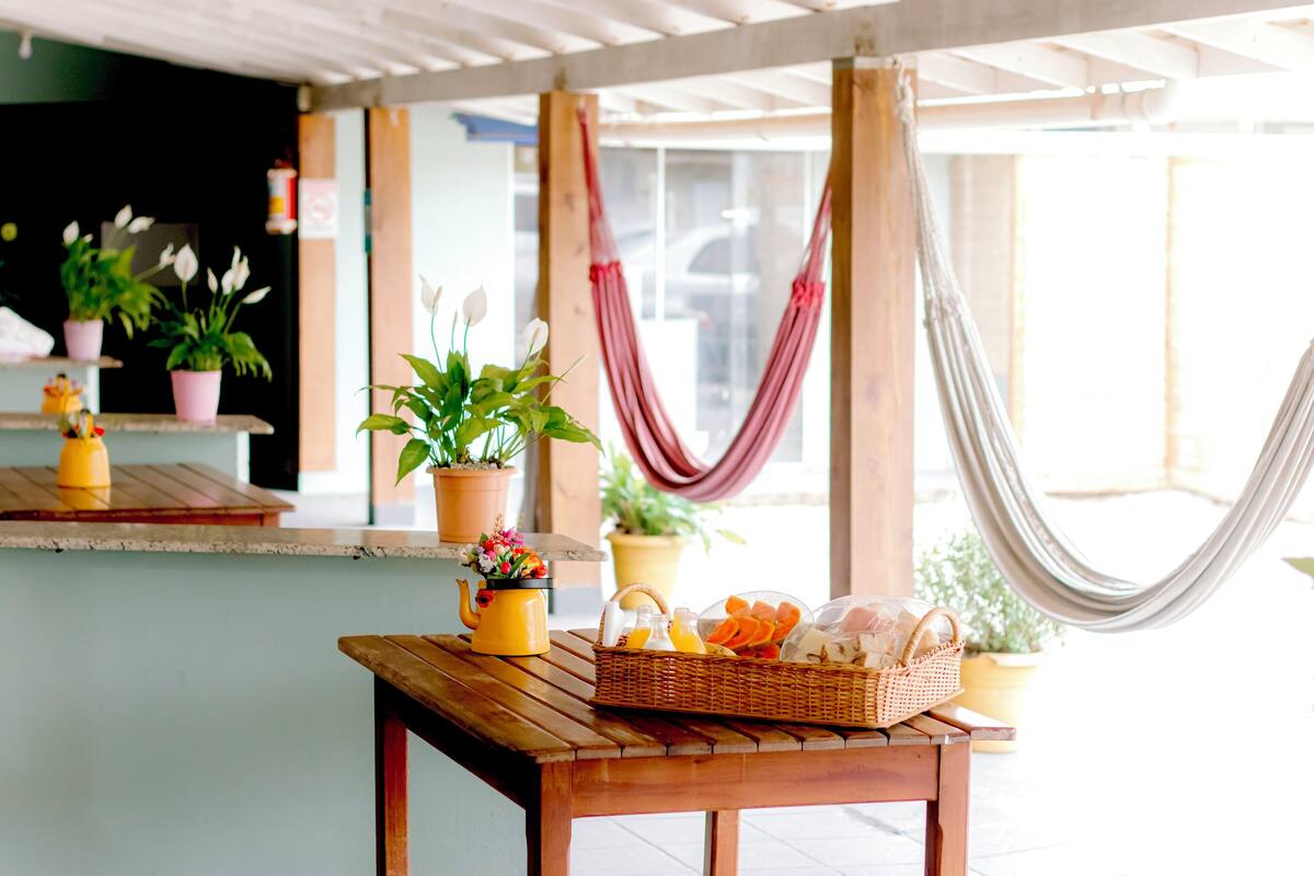 Une table en bois, sur laquelle se trouve un panier de fruits et une théière jaune, entourée de plantes en pot et d’un hamac.