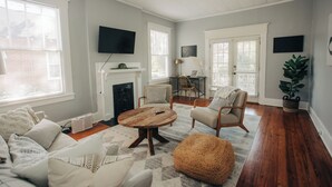 A living room with a round wooden coffee table, a sofa, and two armchairs. There is a fireplace, a television mounted on the wall, and a large window with blinds. - Modern One bedroom near downtown and Augusta National (Augusta)