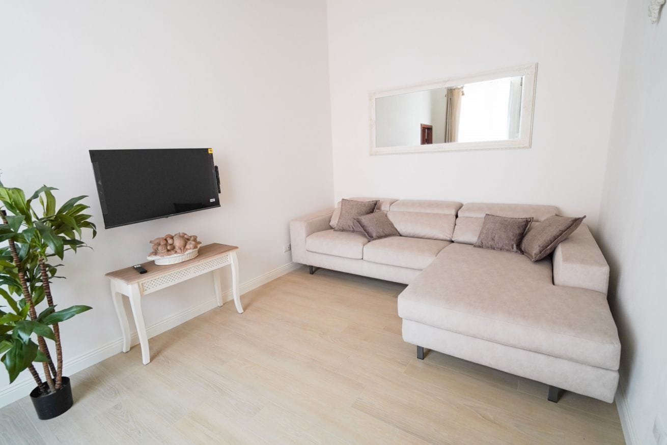 Ein Wohnzimmer mit einem beigefarbenen Sofa, einem Flachbildfernseher, einem kleinen Tisch mit Dekoration und einer Zimmerpflanze.