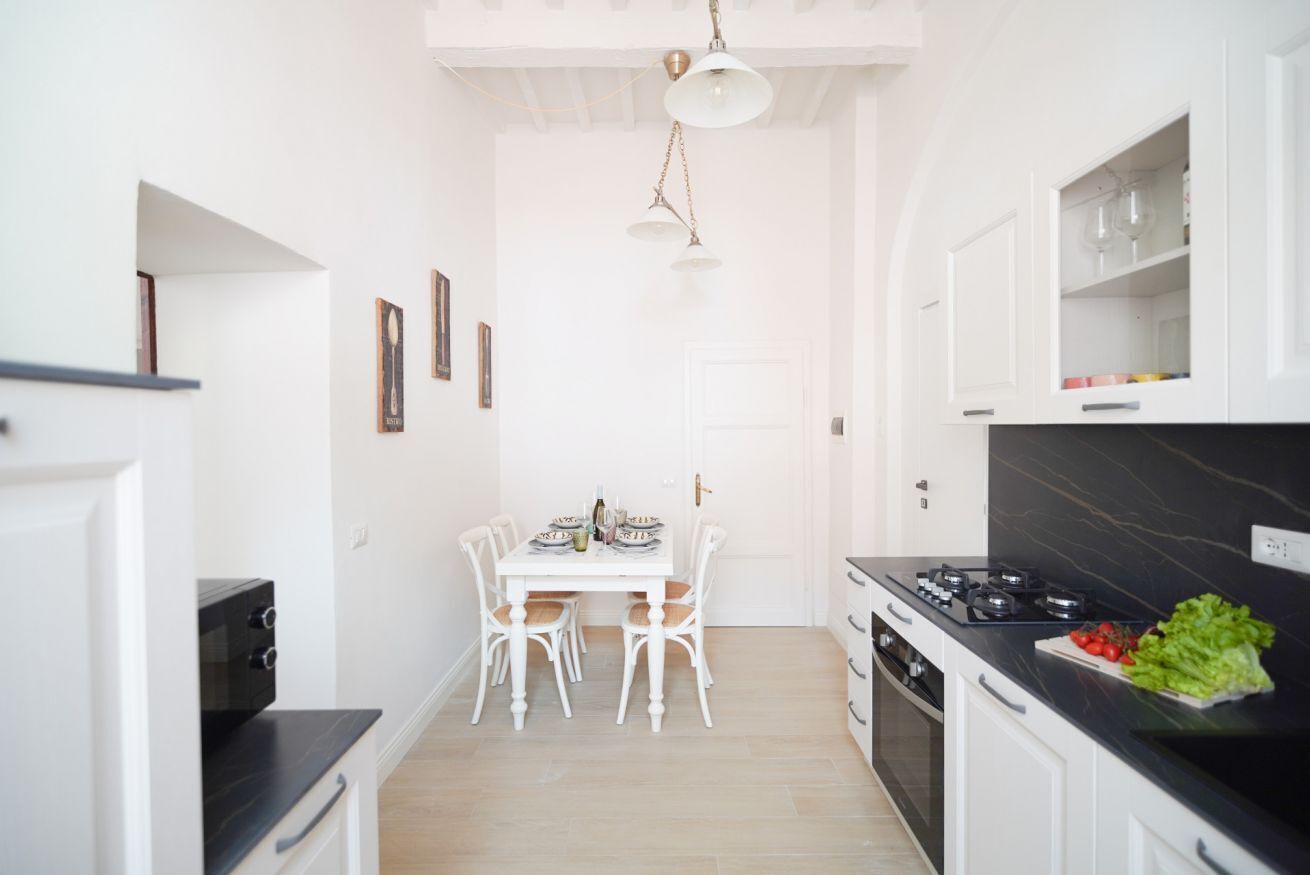 Una cocina compacta con armarios blancos, una mesa de comedor con sillas, una encimera negra y una lámpara colgante.