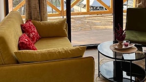 A cozy room with a yellow sofa, a small round table, and a large window offering a view of a snowy landscape and a wooden deck outside. - A frame cabins in the heart of a national park! (Rakovac)
