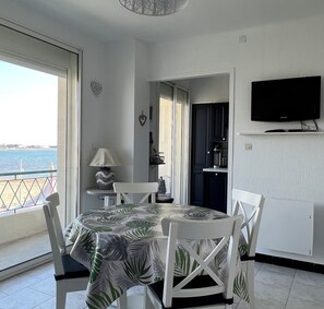 A dining area with a table covered in a tropical leaf tablecloth, white chairs, a lamp, and a view of the sea through a large window. - La Croix du Sud 2 – T2 4 pers proche plage (Le Grau-du-Roi)