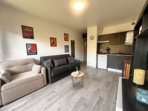 A living room with a sofa, a coffee table, and a kitchen area with cabinets and a sink. - Les Terrasses de l'Espiguette - studio rénové terrasse plage à pied (Le Grau-du-Roi)