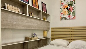 Un dormitorio con cabecera de madera, cama con ropa blanca y estante con varios objetos, incluyendo una placa con nombre, un adorno y una foto enmarcada.