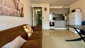Una sala de estar con un sofá marrón, una almohada colorida y una almohada blanca. Se observa una cocina al fondo, con un refrigerador, microondas y estufa.