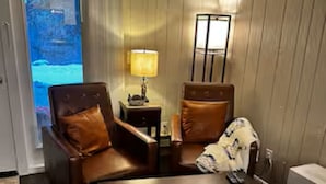 Une chambre d’hôtel avec un mur lambrissé en bois, une porte-fenêtre coulissante donnant sur un balcon, une petite table avec une lampe, deux fauteuils en cuir et un lit recouvert d’une couette blanche.