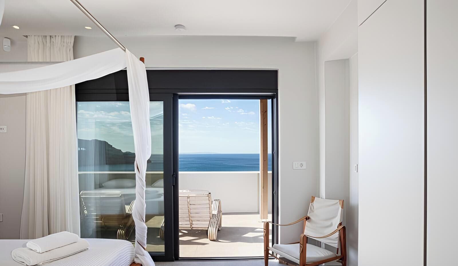 A modern hotel room with a large balcony, a bed with white linens, and a view of the sea.