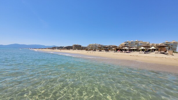 A beach with clear water, a sandy shore, and buildings in the background. - Canet-en-Roussillon T2 Beachfront with Parking - Perfect for 4 (Canet-en-Roussillon)