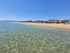 Una platja amb aigua cristal·lina, sorra daurada i edificis al fons.