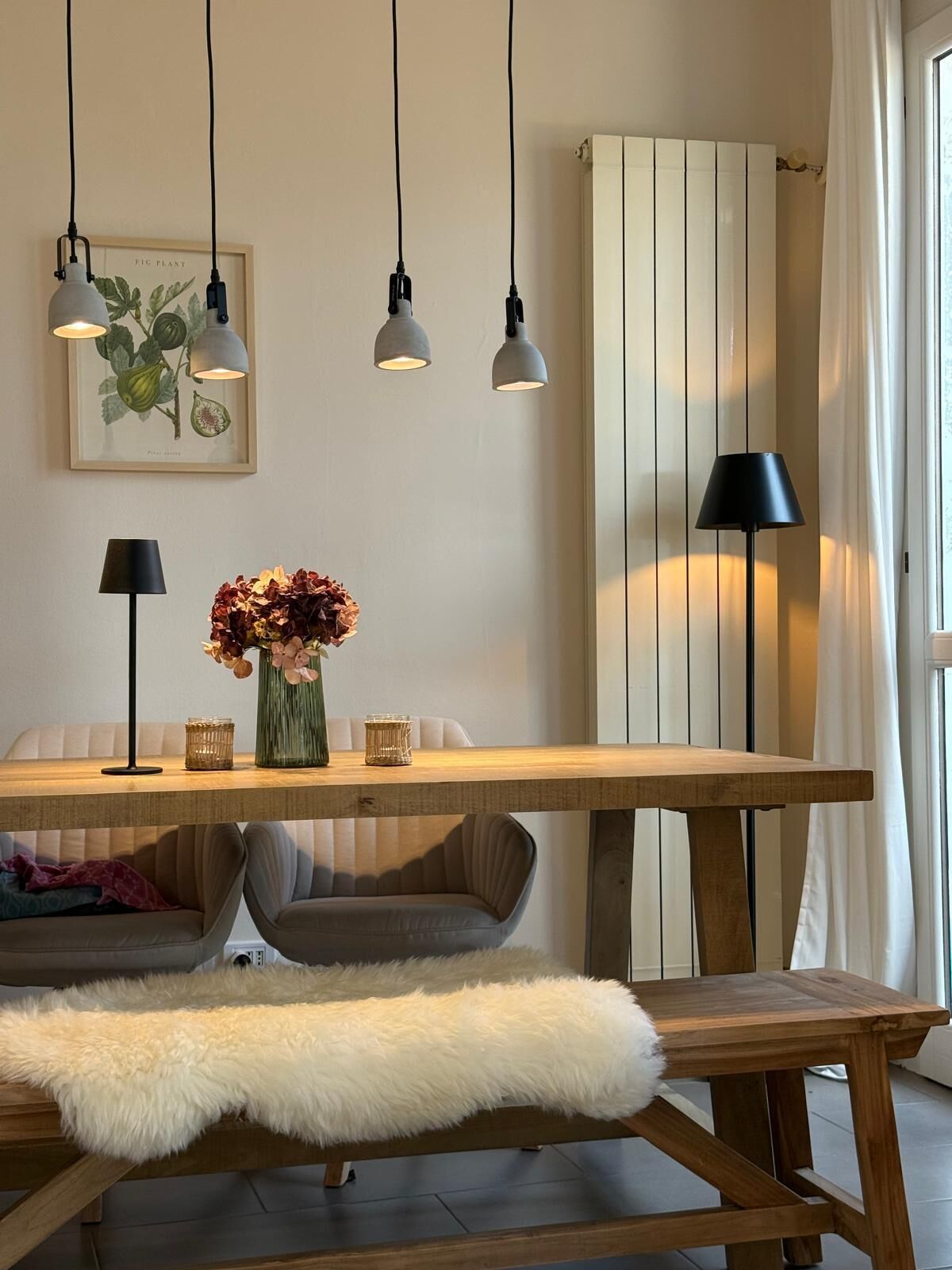 Ein Essbereich mit einem Holztisch, zwei Stühlen, einer Bank mit weißem Fellüberwurf, hängenden Pendelleuchten, einem gerahmten Pflanzenmotiv und einem Heizkörper.