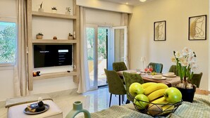 Uma sala de estar moderna com televisão de tela plana, um espaço de refeições com mesa posta para dois e uma grande janela com cortinas.