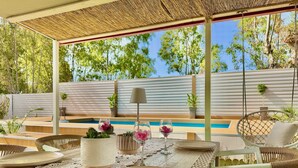 Um pátio ao ar livre com mesa posta para refeições, rodeado por cadeiras de vime e uma cadeira suspensa, com uma piscina ao fundo.